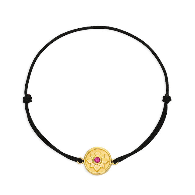 A sterling silver bracelet plated in 18k yellow gold featuring a lotus flower design with a red gemstone at the center, displayed on a black cord.
