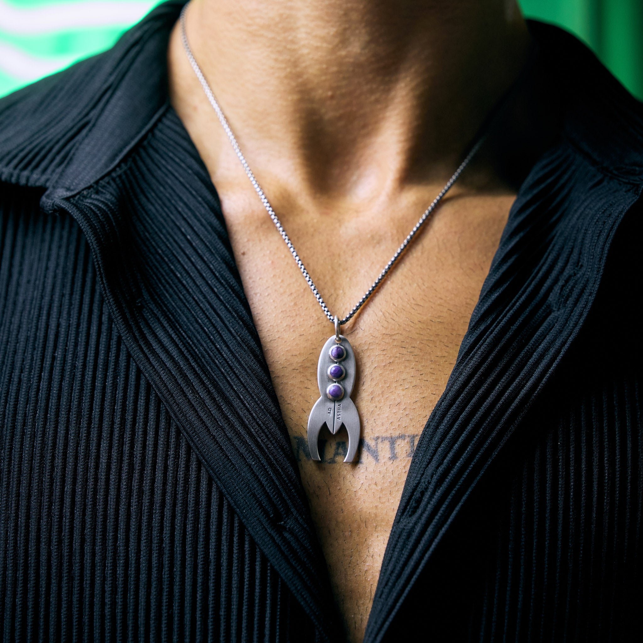 Male model wearing a necklace with a unique rocket pendant, set against a blurred green background