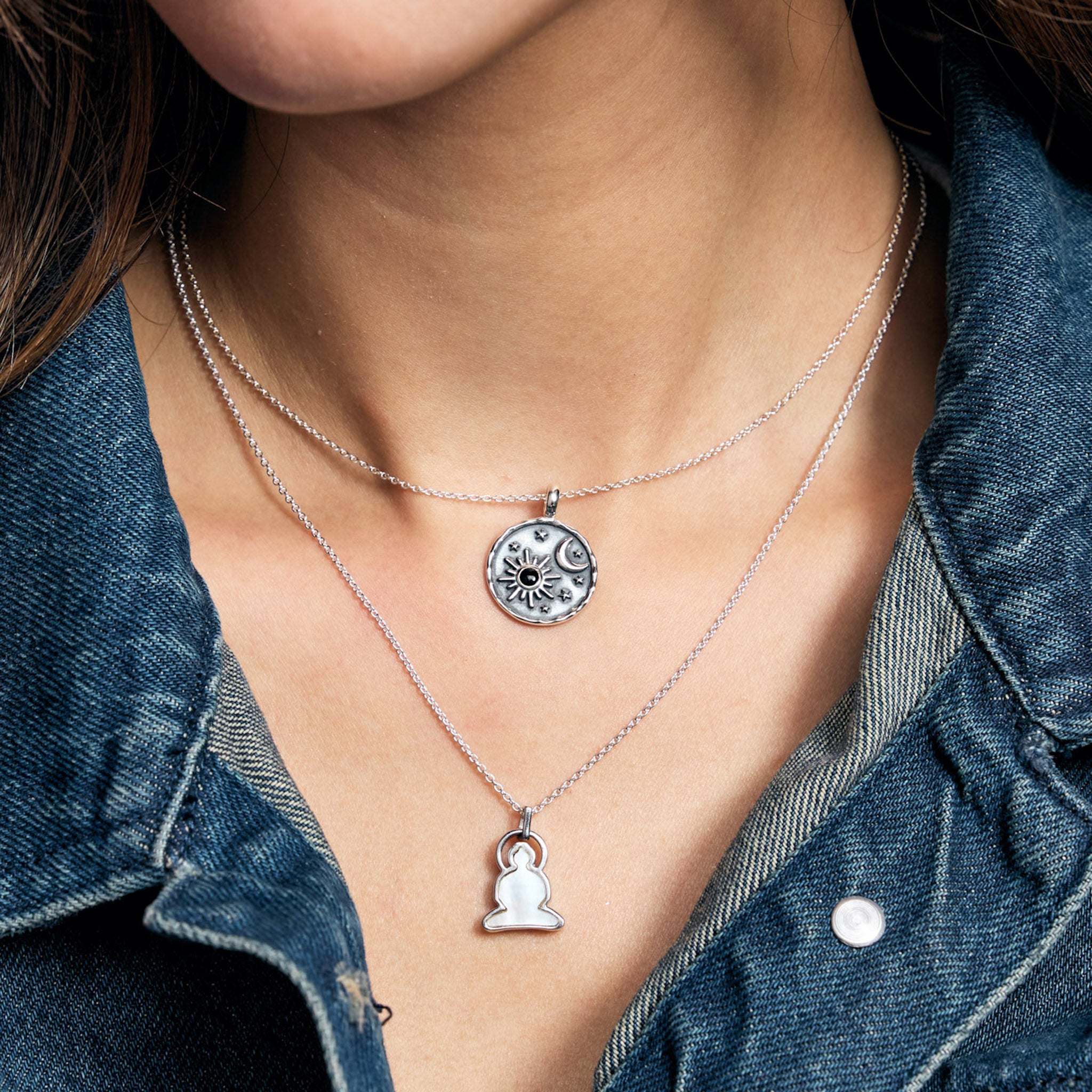 Close-up of a person wearing two silver necklaces with pendants, one featuring a Buddha figure and the other a constellation design, against a denim background.