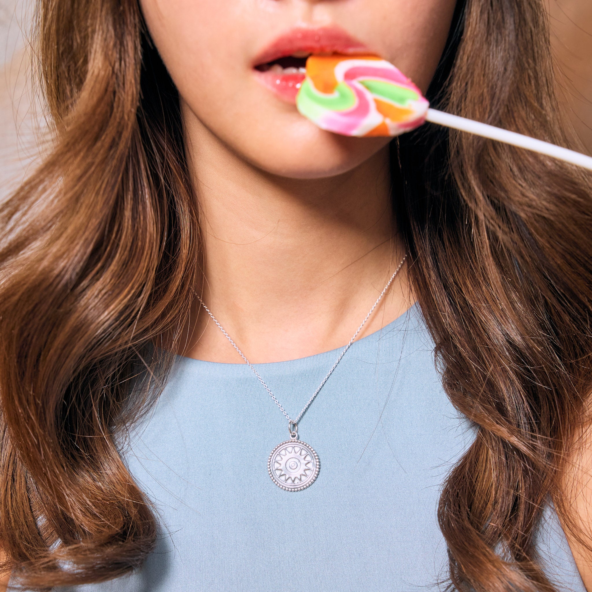 Close-up of a female model with long brown hair wearing a blue top and a sun silver necklace. She is holding a colorful lollipop.