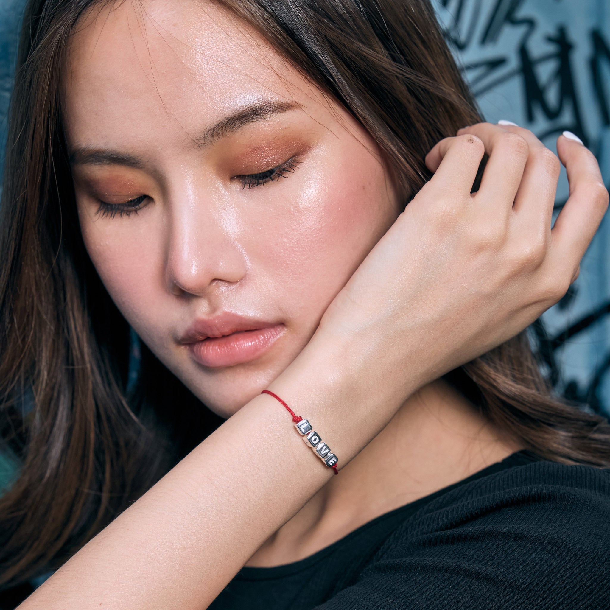 Woman wearing a bracelet with a visible love charm against a blue graffiti wall.